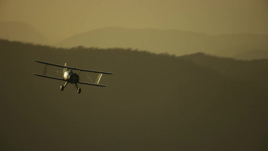A biplane flies in a hazy yellow sky with mountains in the distance