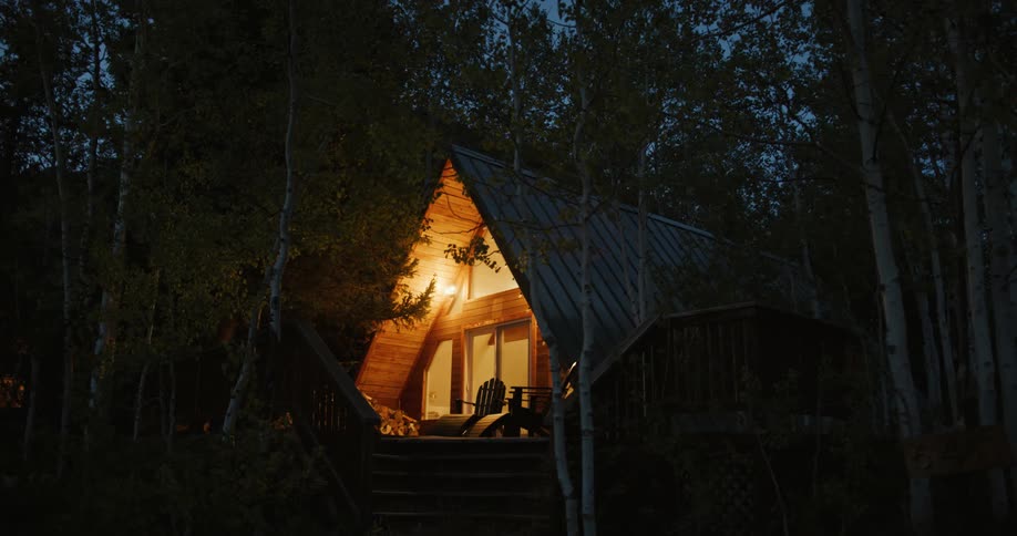 A light comes on the front porch of an a frame house in the woods