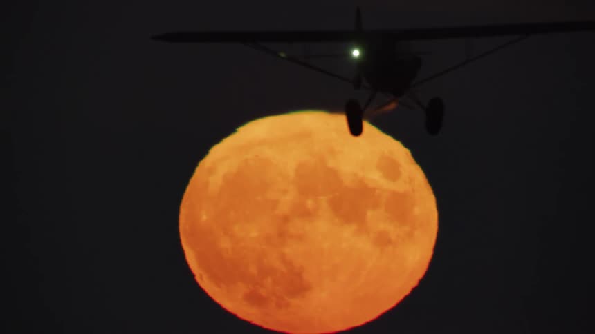 Small flying plane crosses the face of the moon and is silhouetted against the sky