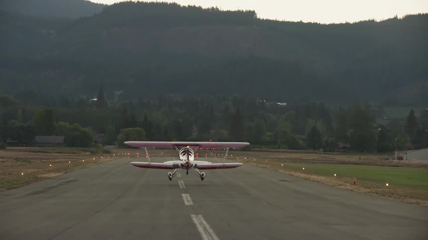 Small biplane takes off down the runway then begins to fly towards the mountain as the camera looks down the runway