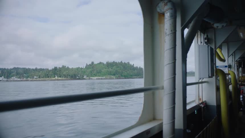 Large boat with open windows cruises across water with view of town and woods on the shore