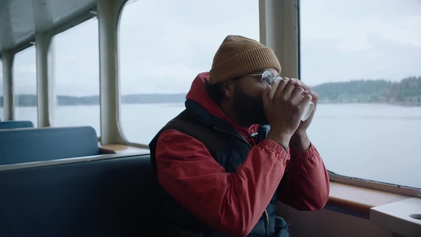 Man sitting inside a ferry crossing a lake takes a sip of a hot beverage