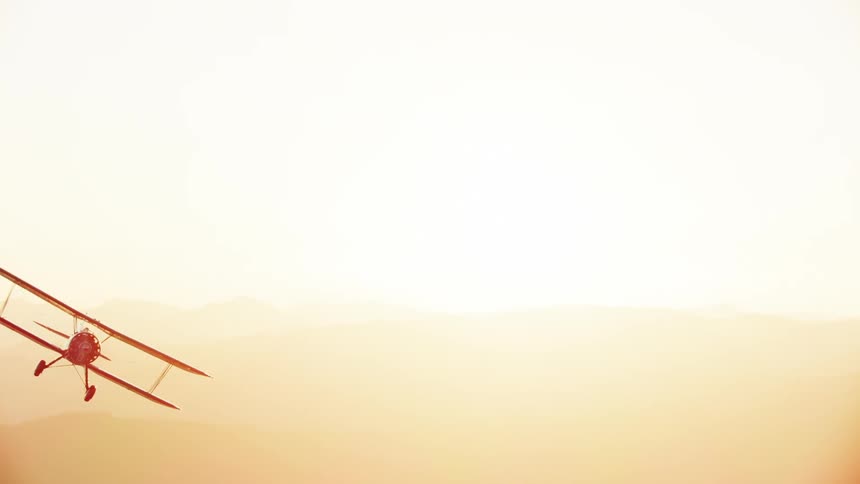 Old red biplane flying in front of the sun setting over the mountains