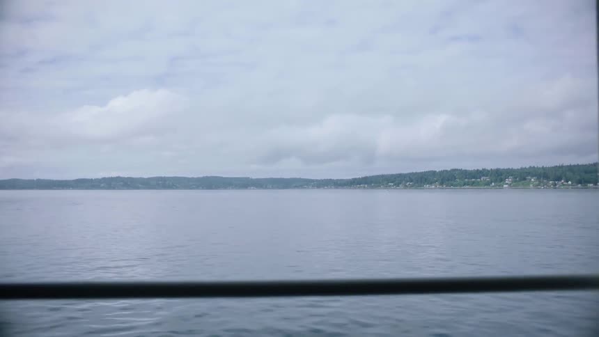 Boat travels on the ocean as land with hills and buildings sit in the distance under a cloudy sky