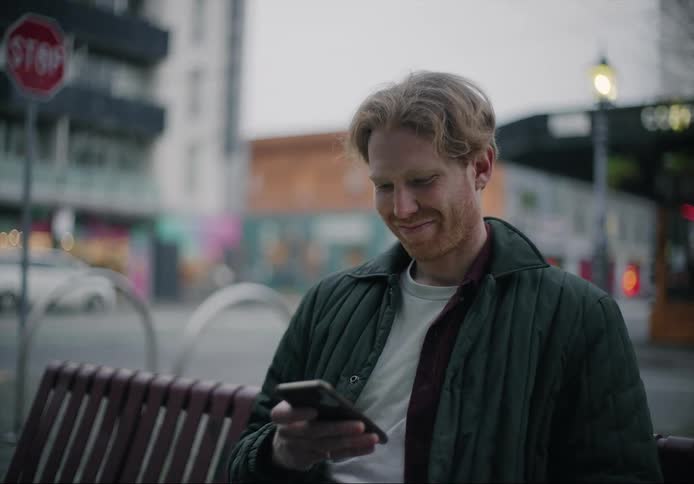 A man is sitting on a bench near a city street looking at his cell phone with a smile on his face