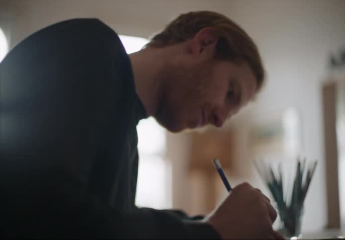 Man with beard and black sweater sits at table writing down in room