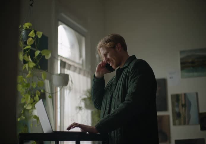 A man is standing in a room typing on a laptop computer when he reaches into his back pocket and takes out his phone and answers it