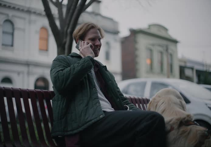 A man is sitting on a bench near a downtown street with his dog and talking on his cell phone