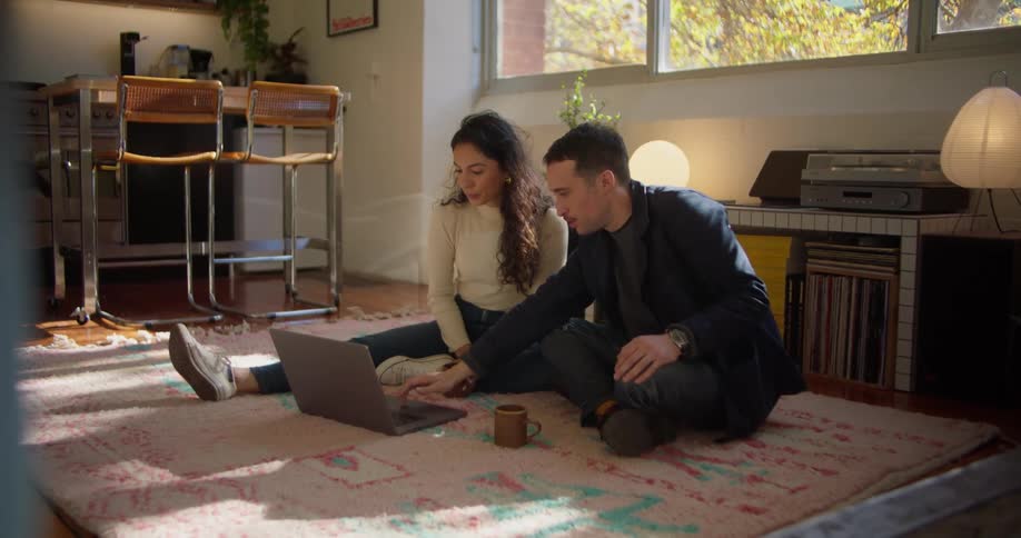 A woman and a man are sitting on a living room floor with a laptop computer and they are talking about what they are seeing on the internet