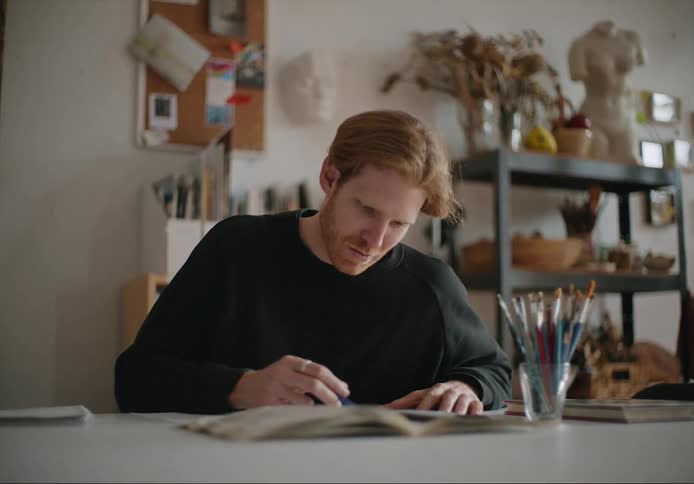 A man stops sketching with a pencil and looks up smiling while sitting in his art studio