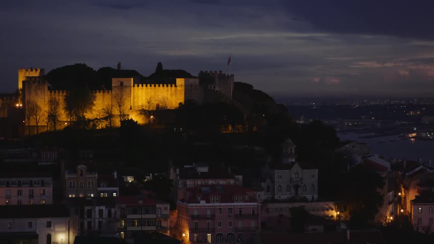 Flag waving over the walls of a castle or fort atop a mountain on a town at nightfall