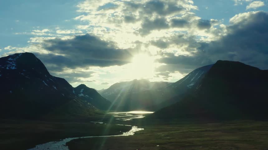 The sun shines and beams down from the clouds on mountain range with patches of snow and a winding stream