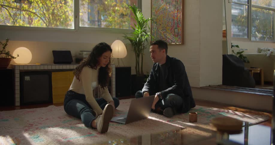 Woman with brown hair white sweatshirt and jeans sits on rug next to man with mustache black jacket and jeans working on laptop in living room at home