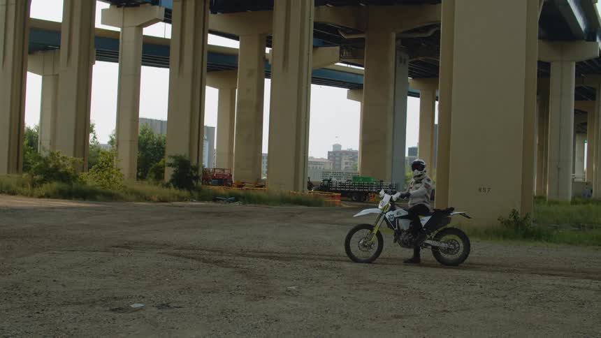 Man wearing protective gear sitting over a parked motocross motorcycle under a highway