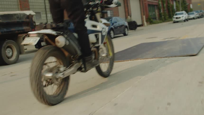 Motorcyclist rides on a city street and lifts the front of the bike while traveling over a large sheet of plywood