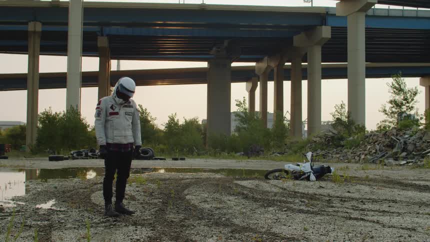A man wearing a motorcycle helmet rotates his neck after crashing his motorcycle on a dirt road in front of a raised highway