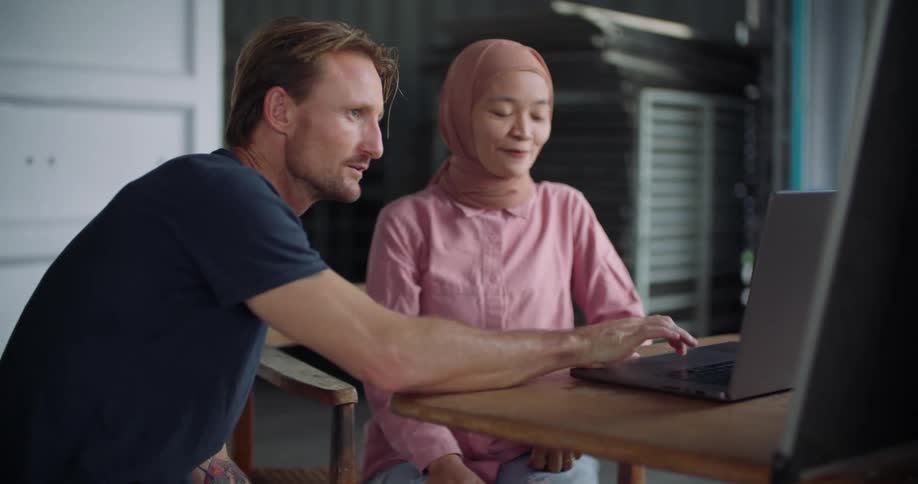 A young woman and a man talk while sharing a laptop at a table indoors during the day