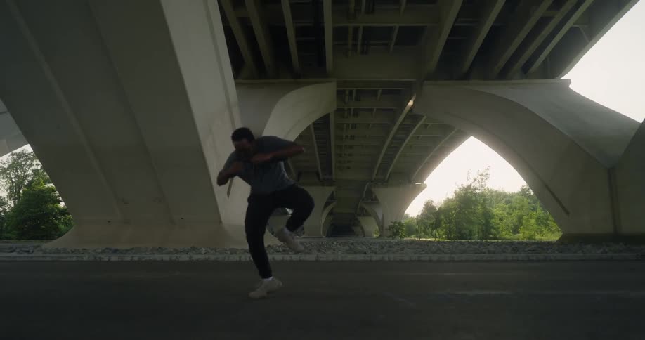 A man does a hip hop style dance under a bridge