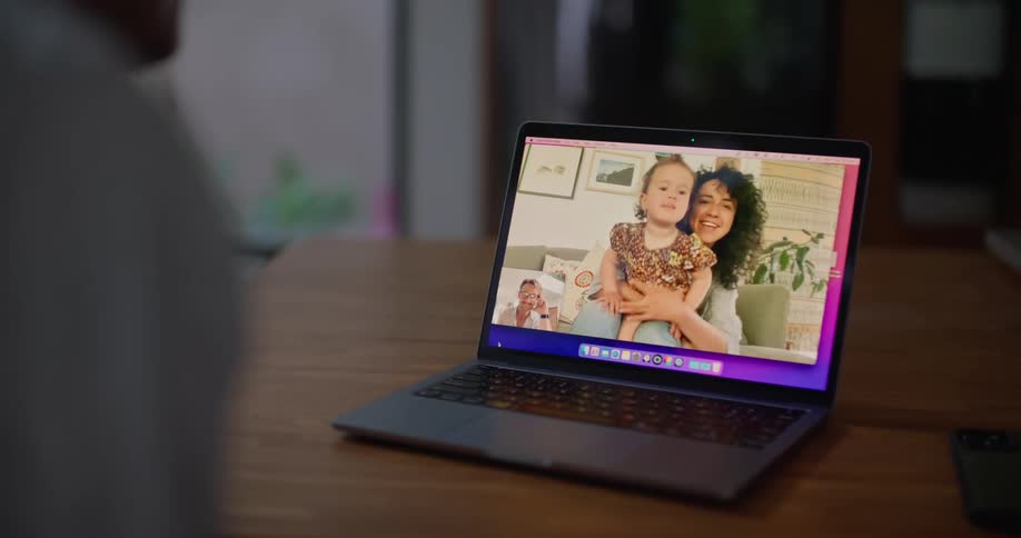 Dad waves at the laptop screen where mom is holding their daughter on her lap and laughing