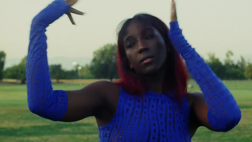 A black girl with red hair and blue gloves is wearing a blue dress dancing with her arms while looking into the camera with a blank expression on her face