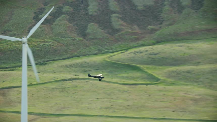 Small airplane flies over a field and past the wind turbines in the backcountry