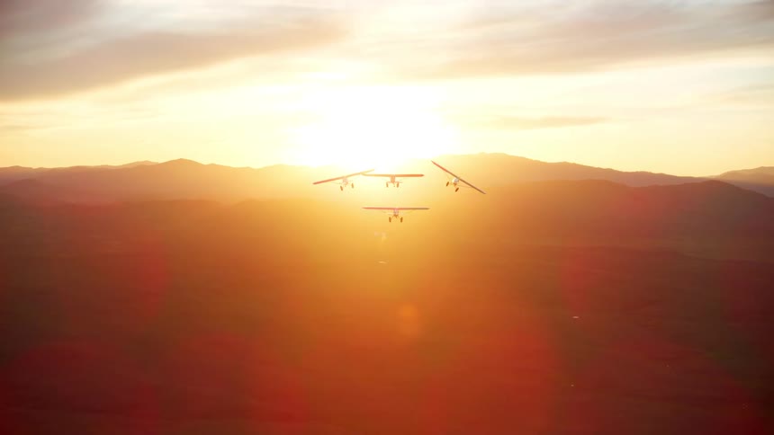 Four small backcountry airplanes are flying in formation and there is a sunset in an orange sky over mountains in the distance