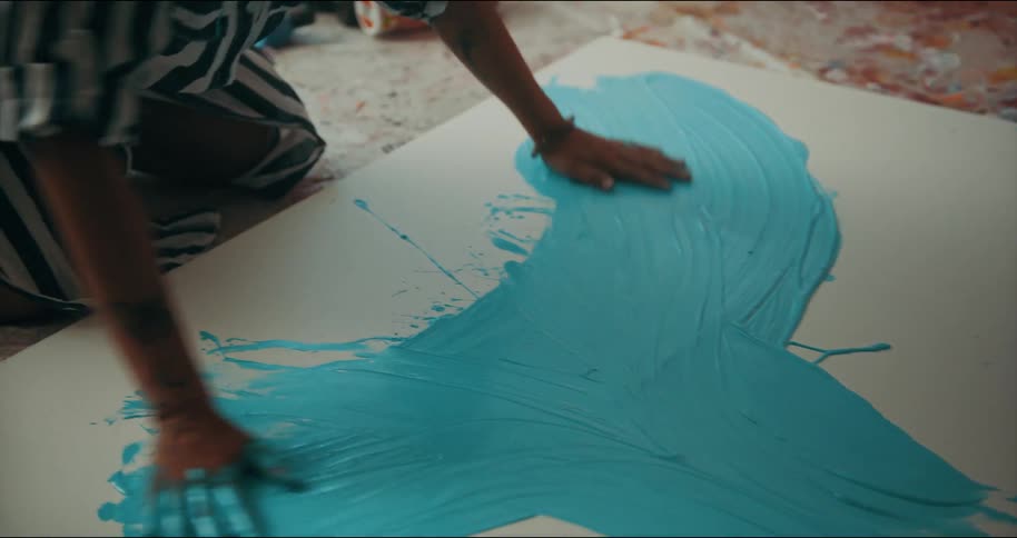 Blonde woman kneeling on the floor of a studio paints with bright colors over canvas using her hands