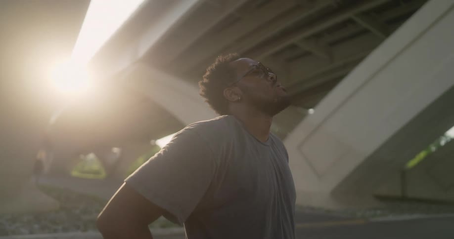 A man is standing below an overpass looking up at the road as the sun shines brightly upon him
