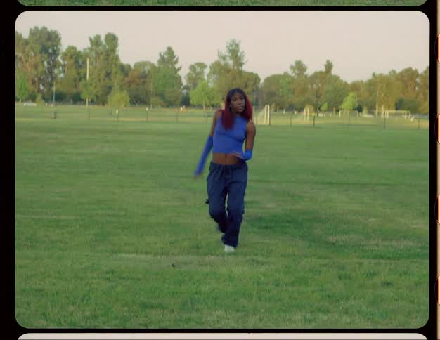 Black woman wearing long blue gloves and a matching blue top moves her arms as she dances in a field near the soccer fields