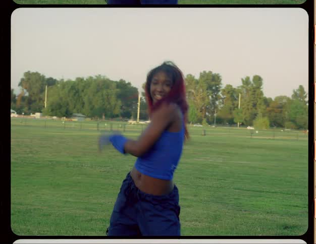 A teenage black girl with long red hair is dancing in a field wearing a blue outfit with blue gloves