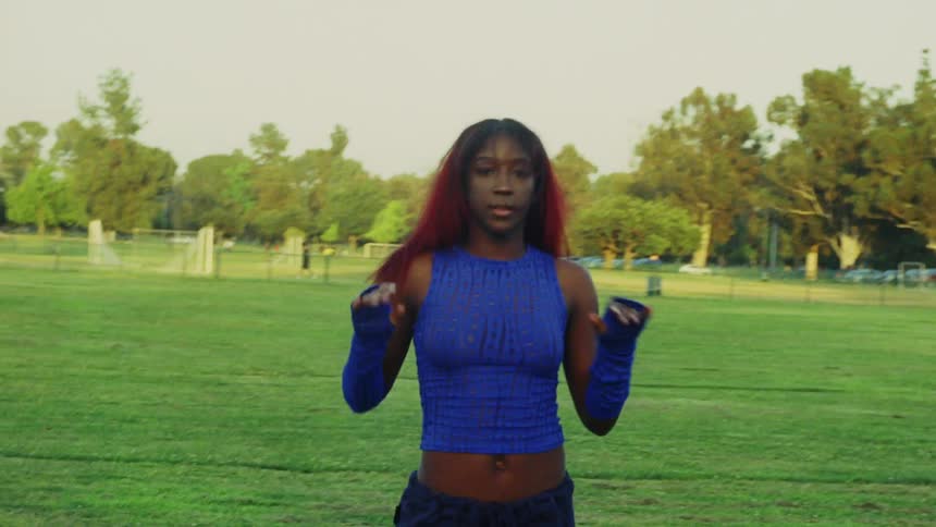 A teenage girl wearing a blue outfit is outside dancing in a field and there are trees in the distance