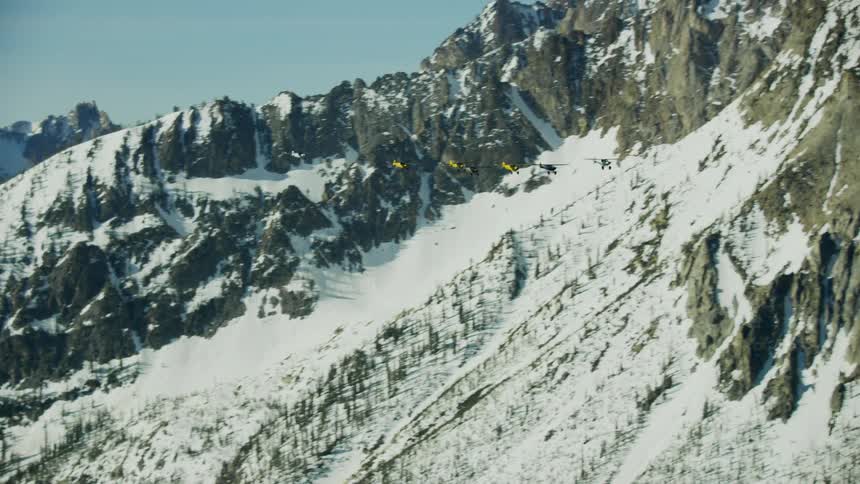 Seven small backcountry airplanes fly over the snowy mountains under a blue sky in the Idaho Backcountry