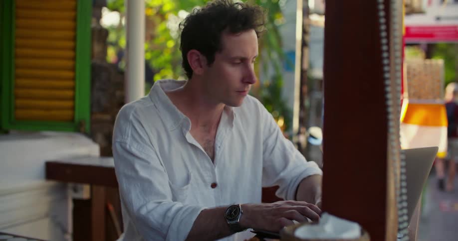 Man sits on porch typing on a laptop as a pedestrian walks away in the background
