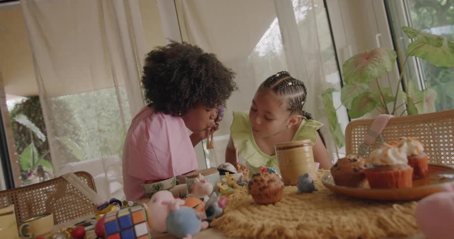 Two young girls are standing near a table with cup cakes playing with a doll and there is a candle lit in a cupcake and one of the girls blows out the candle