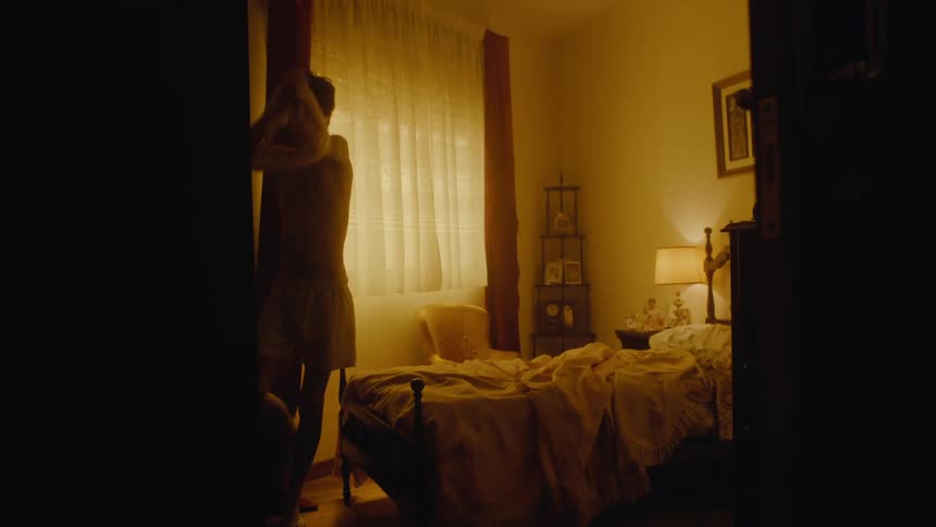 A teenage boy is standing in a bedroom wearing boxer shorts and putting on a tank top shirt as he walks out of the room