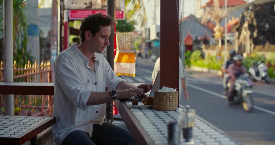 A man is sitting at an outdoor cafe typing on a laptop computer as traffic passes by on the street