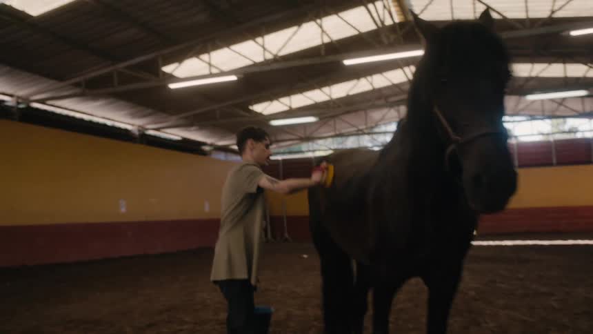 A man is standing inside a large building holding a brush and a bucket as he grooms a horse