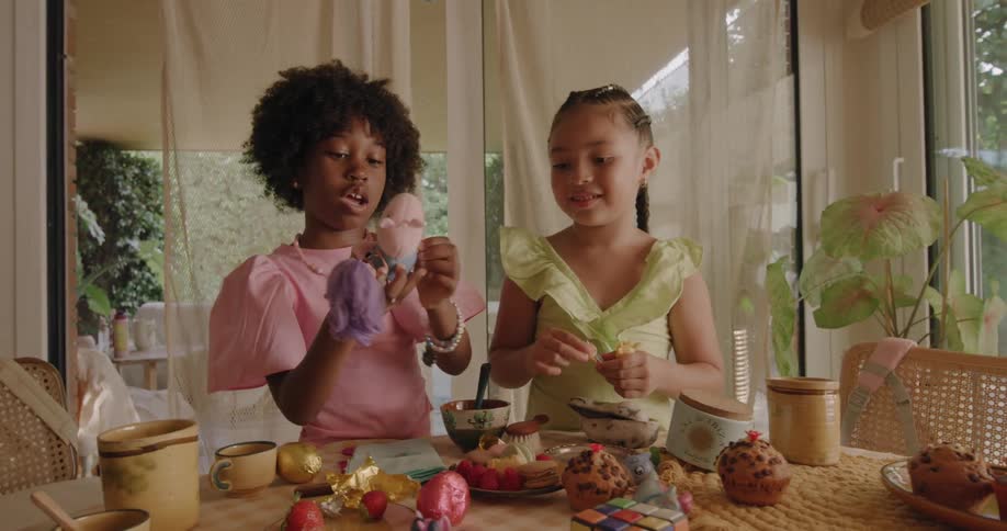 Two girls play with dolls at table with muffins and other desserts