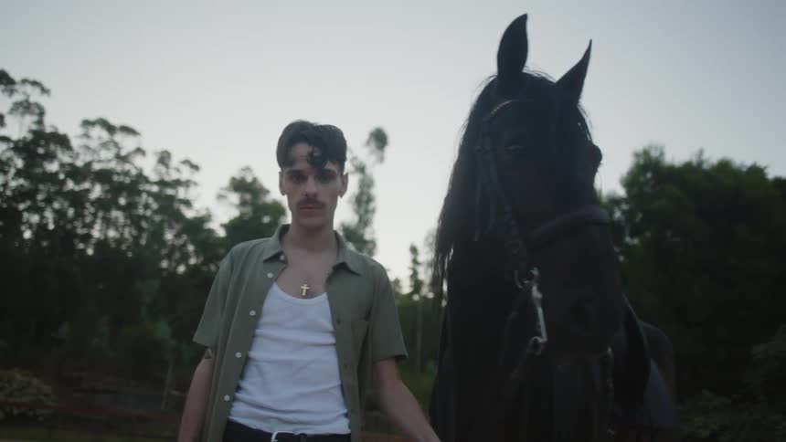 Man with mustache cross necklace and white green shirt stands next to dark horse in front of forest