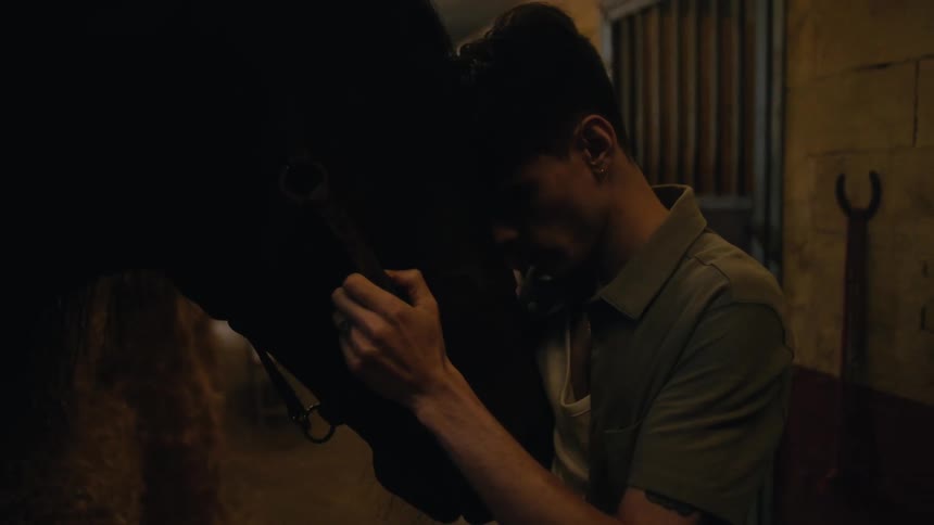 A man is standing in a barn with his horse and leaning his forehead on the horses head as he holds it