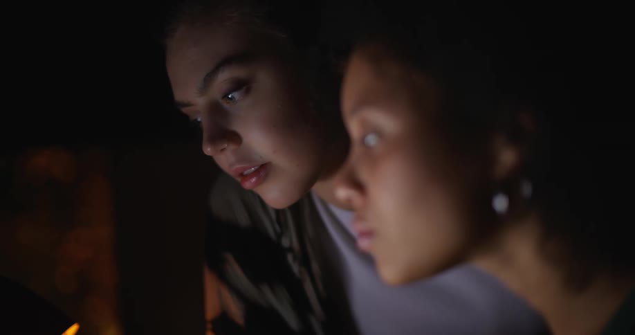 The faces of two young women are illuminated by a laptop screen that they are watching in a dark room