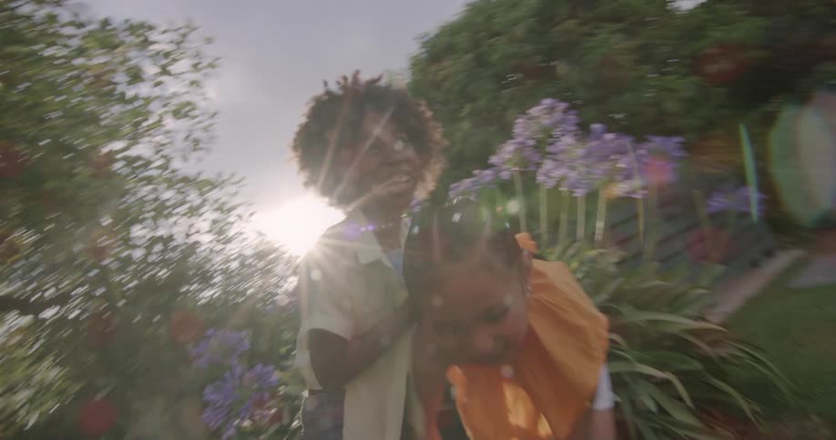 Two young girls are standing outside in a park near flowers laughing and talking as the sun shines brightly in the sky