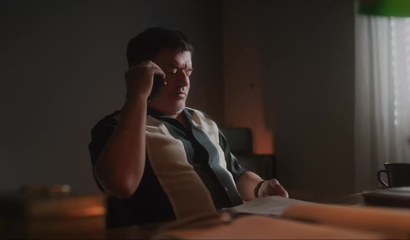 A man sitting at a desk leans back as he talks on the phone while reviewing a document