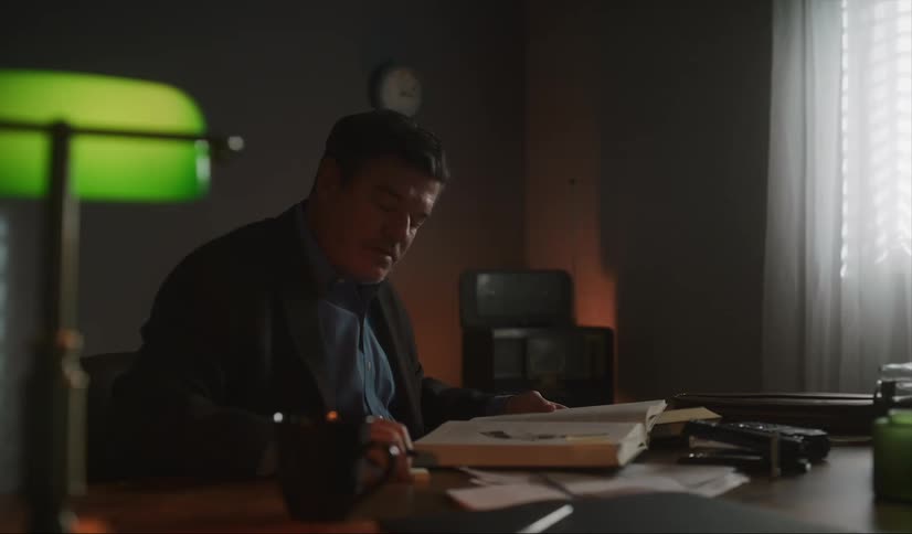 A man sits at his desk while reading a book and then puts it down to make a note on one of its pages