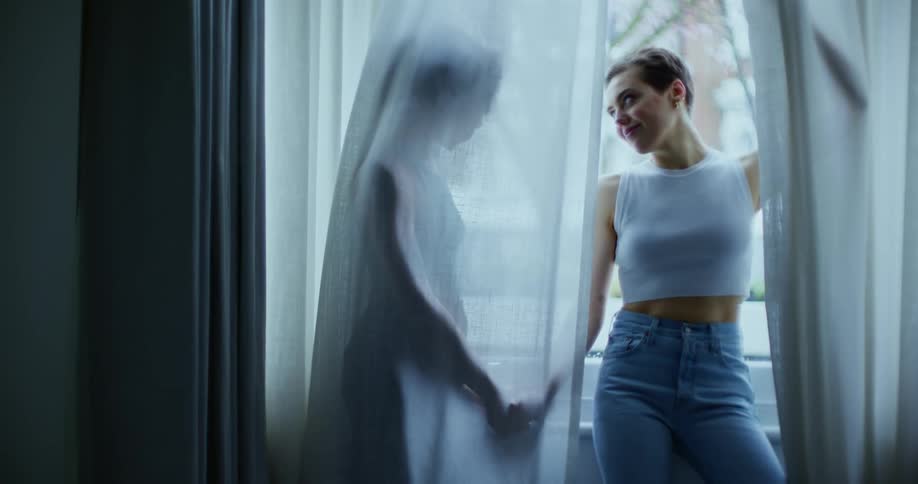 Two women holds hands while posing behind curtains by a large window