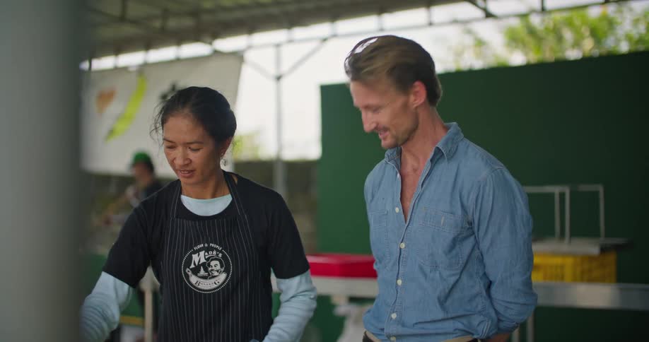 A man is standing next to a woman wearing plastic gloves and moving food on a tray as they talk
