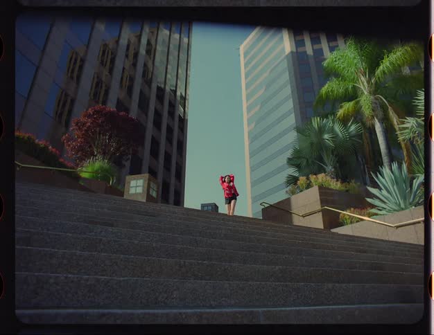 A woman dancing at the top of city plaza steps near palm trees in planters and skyscrapers