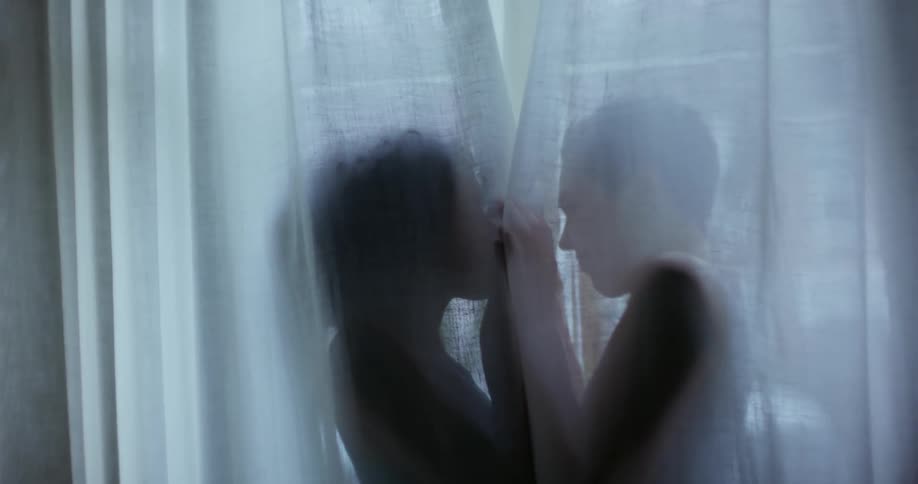 Two women laugh while holding hands as they stand behind a sheer curtain