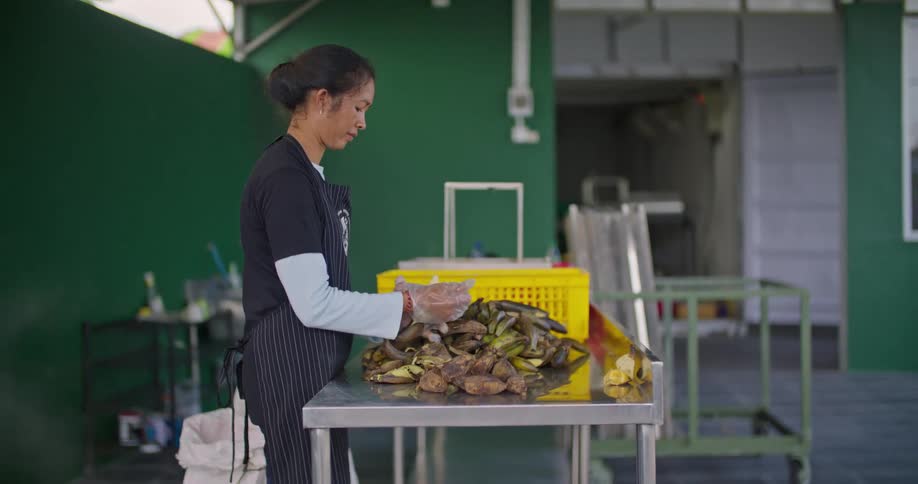 Woman stands at metal table peeling food as smoke drifts by