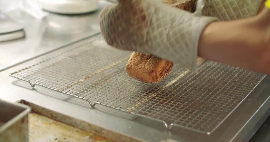 A baker takes fresh loaves of bread out of the molds and places them on a rack
